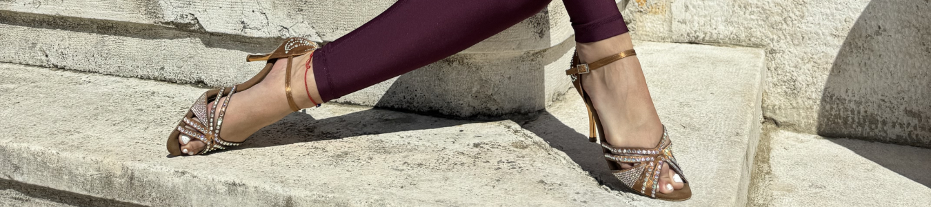 Eine Person in Riemchensandalen mit Verzierung und kastanienbraunen Leggings sitzt im Sonnenlicht auf Steinstufen.