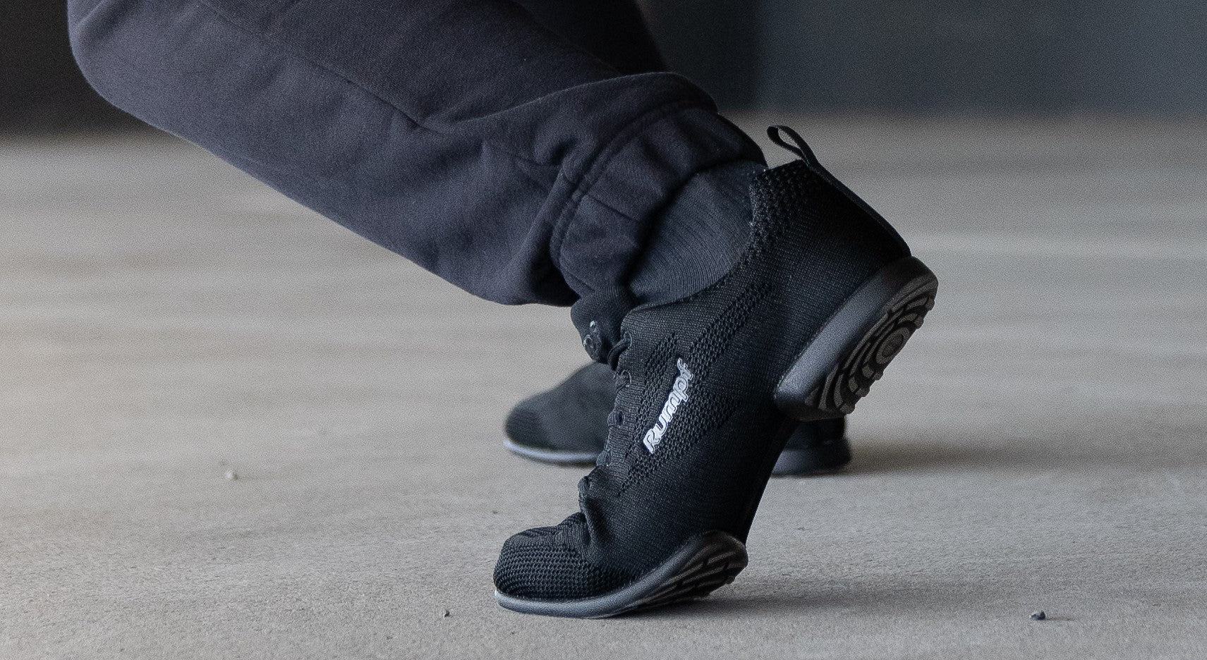 Eine Person in schwarzen Sportschuhen und dunklen Hosen balanciert auf ihren Zehen auf einer Betonoberfläche.