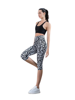 Frau in Lisminas Schwarz & Weiss 941 Capri-Leggings und schwarzem Sport-BH, balanciert auf einem Bein vor einem schlichten Hintergrund.