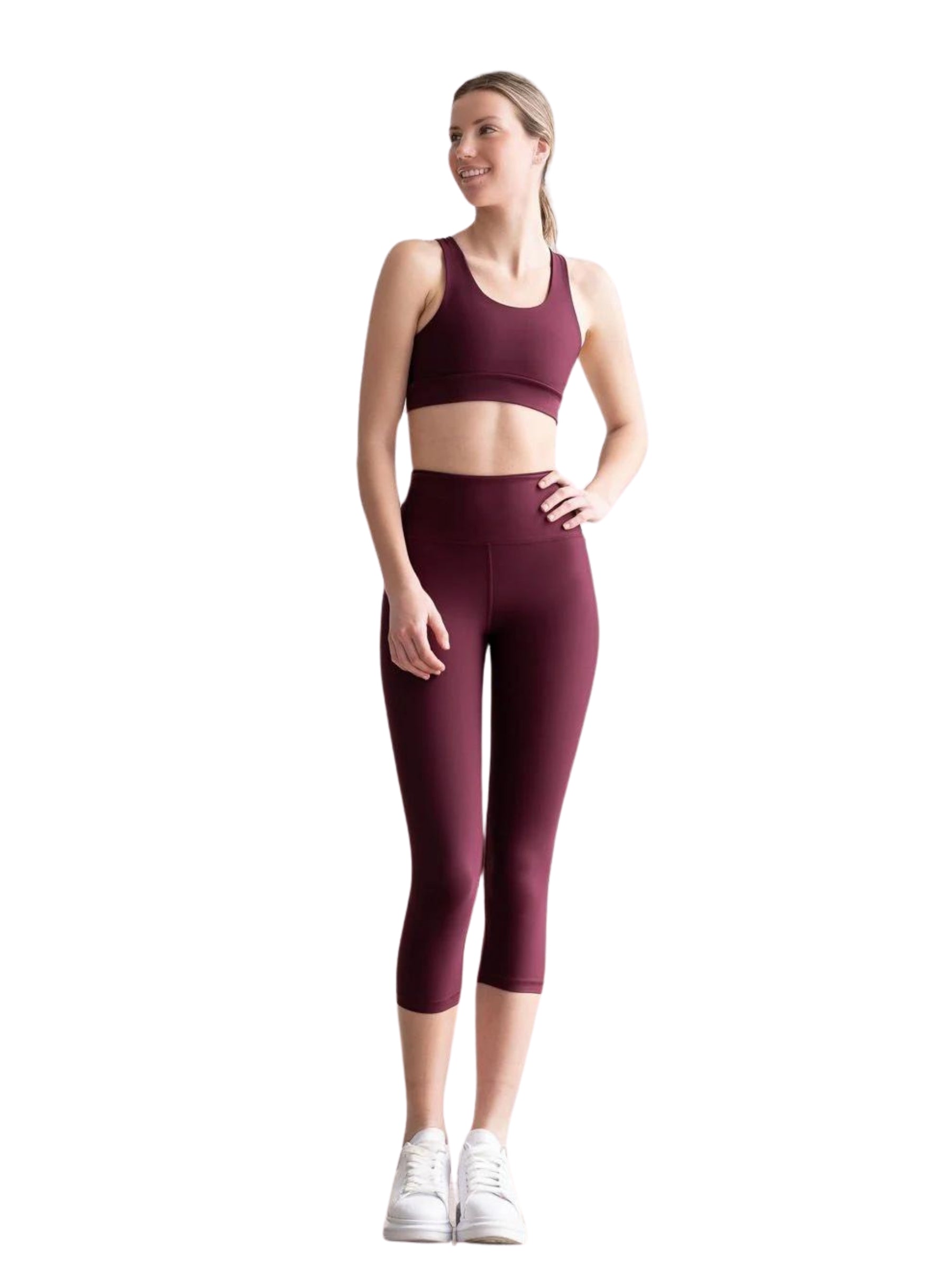 Frau in Capri-Leggings „Lismina Bordeaux“ und kastanienbraunem Sport-BH lächelt in weißen Turnschuhen auf weißem Hintergrund.