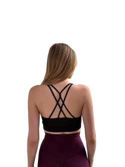 Eine Frau trägt einen Lismina 247 Fit Sport-BH in Schwarz, mit lila Leggings und überkreuztem Rücken und blickt abgewandt.