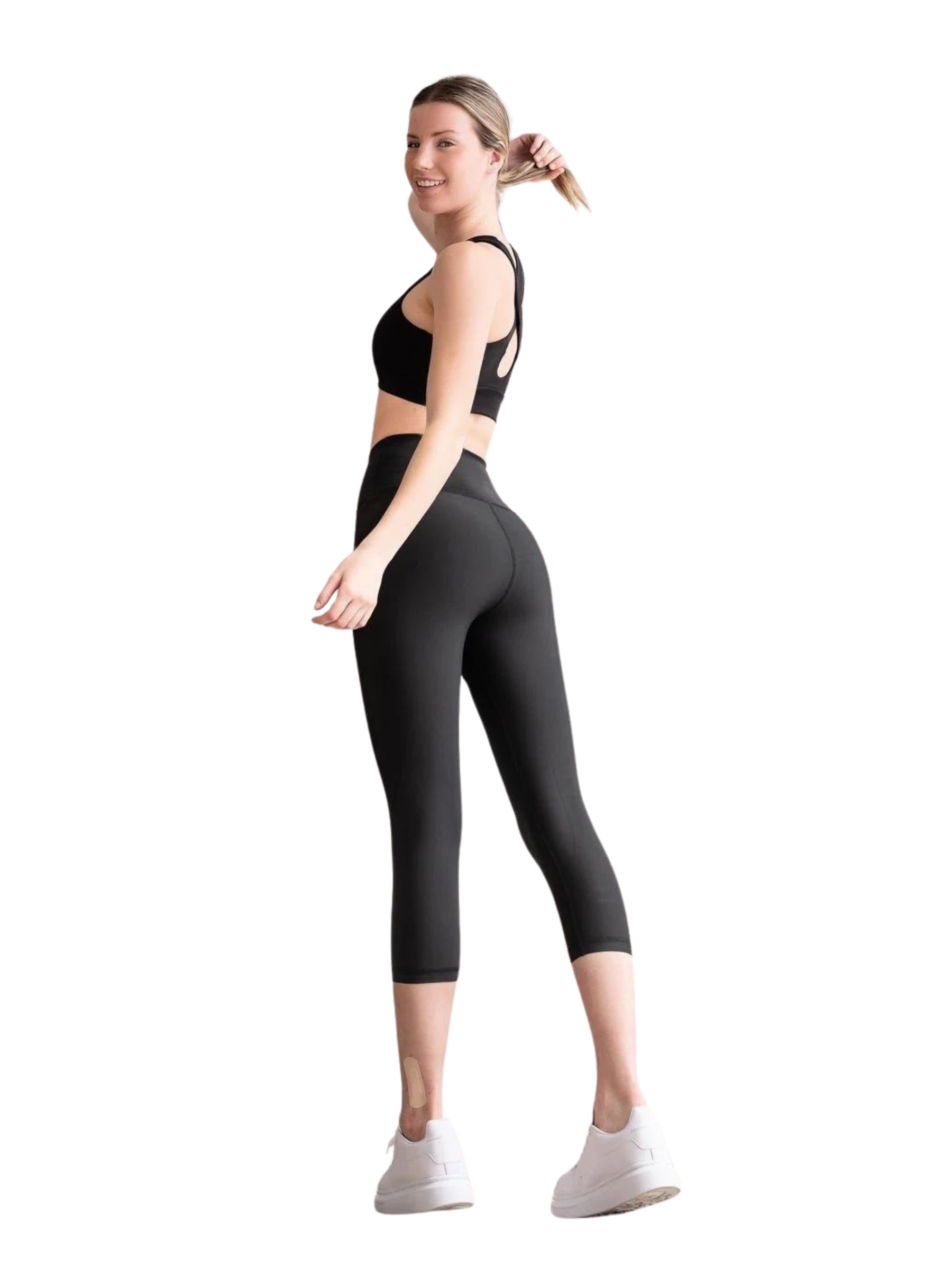 Frau in Lisminas 4306 Capri Leggings Schwarz, weißen Turnschuhen, mit dem Rücken nach hinten gerichtet, lächelt und hält ihren Pferdeschwanz.