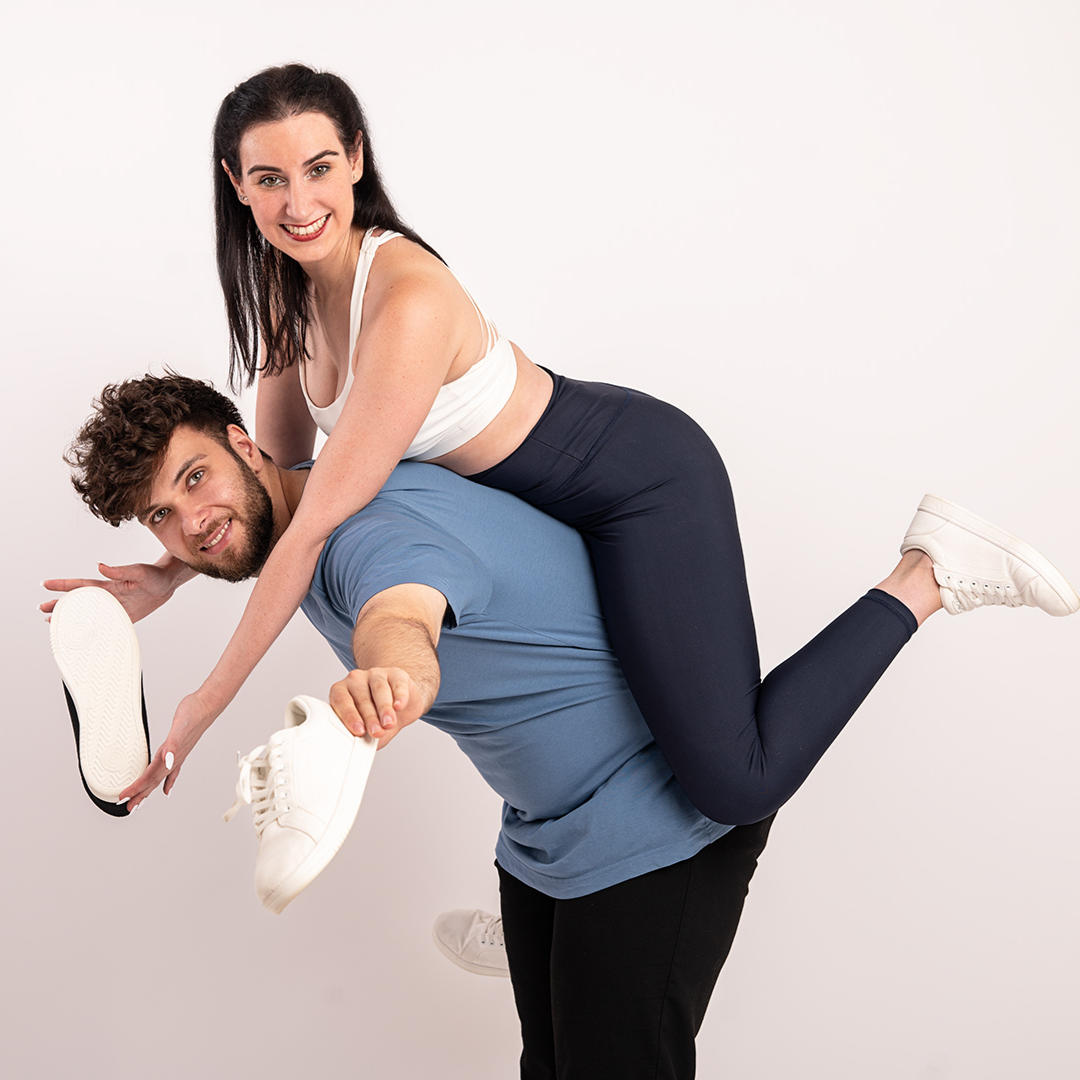 Ein Mann hält spielerisch eine Frau auf seinem Rücken, beide lächeln und haben weiße Turnschuhe in der Hand, vor einem weißen Hintergrund.