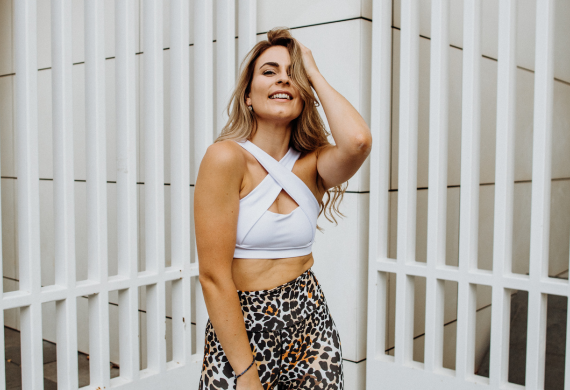 Eine lächelnde Frau in einem weißen Sportoberteil und Leggings mit Leopardenmuster steht vor einem weißen Zaun.