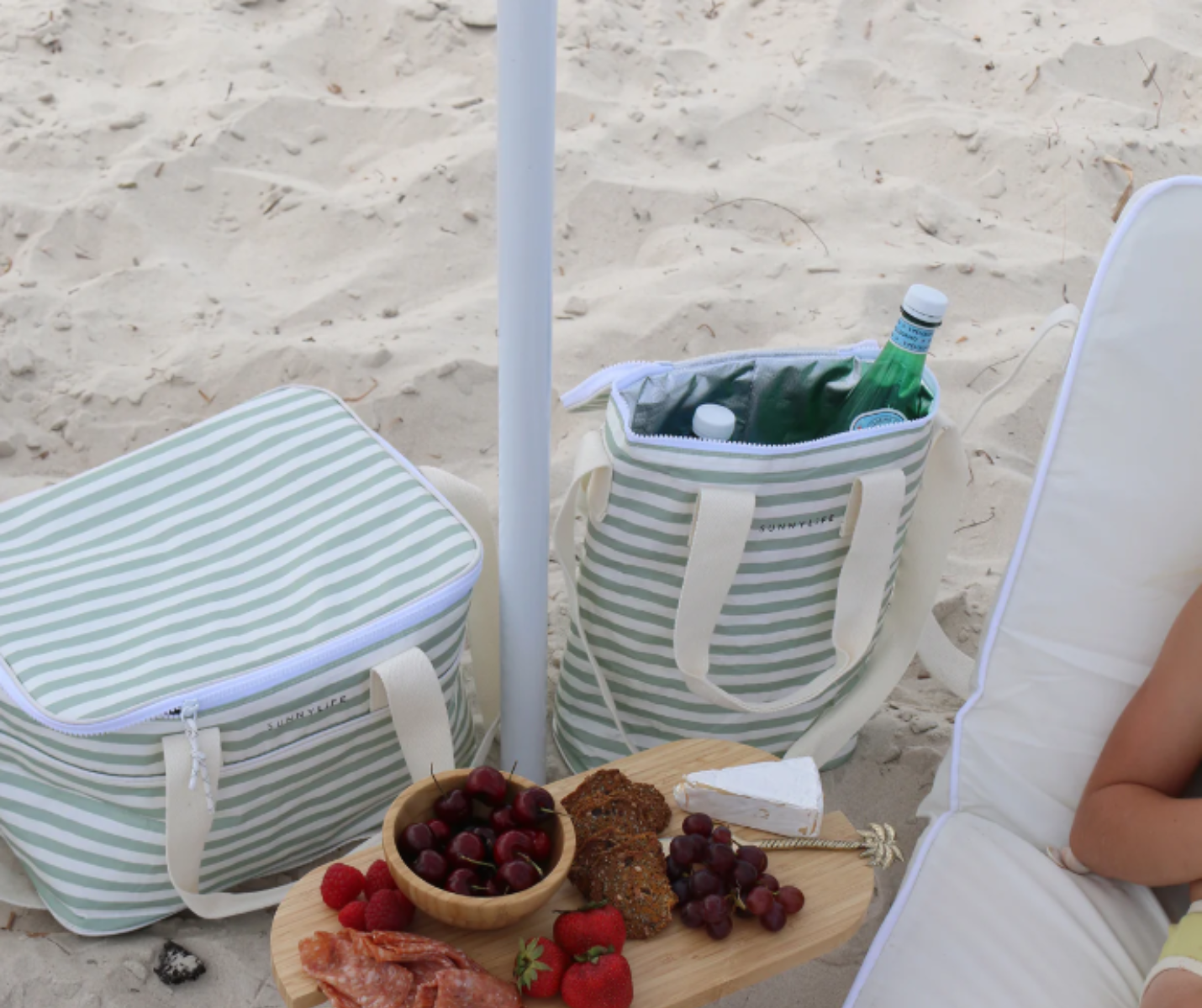 Zwei Xinnomix-Kühltaschen und ein Snackbrett liegen neben einer Person, die sich unter einem Sonnenschirm entspannt.