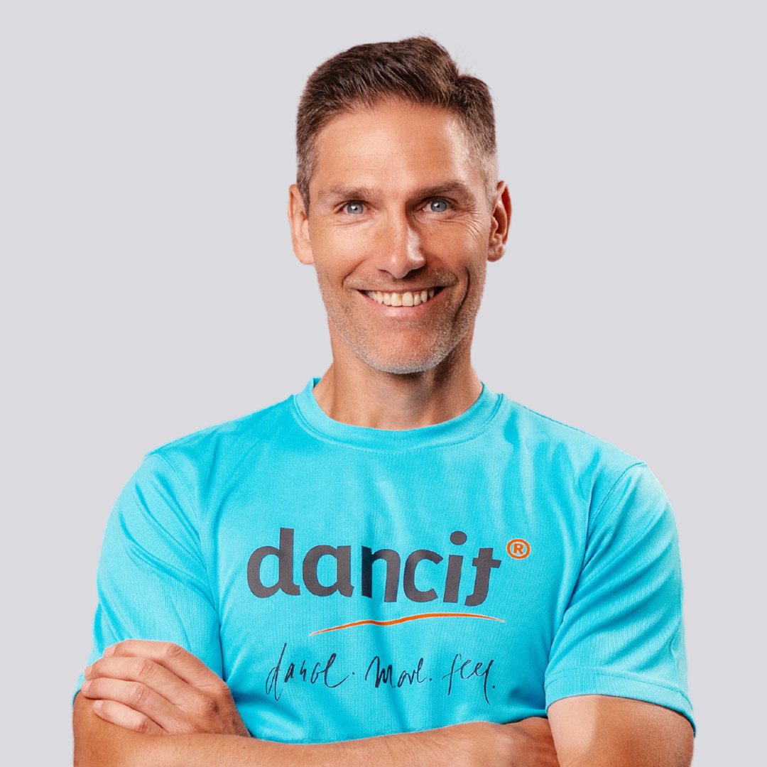 Ein lächelnder Mann mit kurzen braunen Haaren in einem blauen Tanzhemd steht mit verschränkten Armen vor einem hellen Hintergrund.