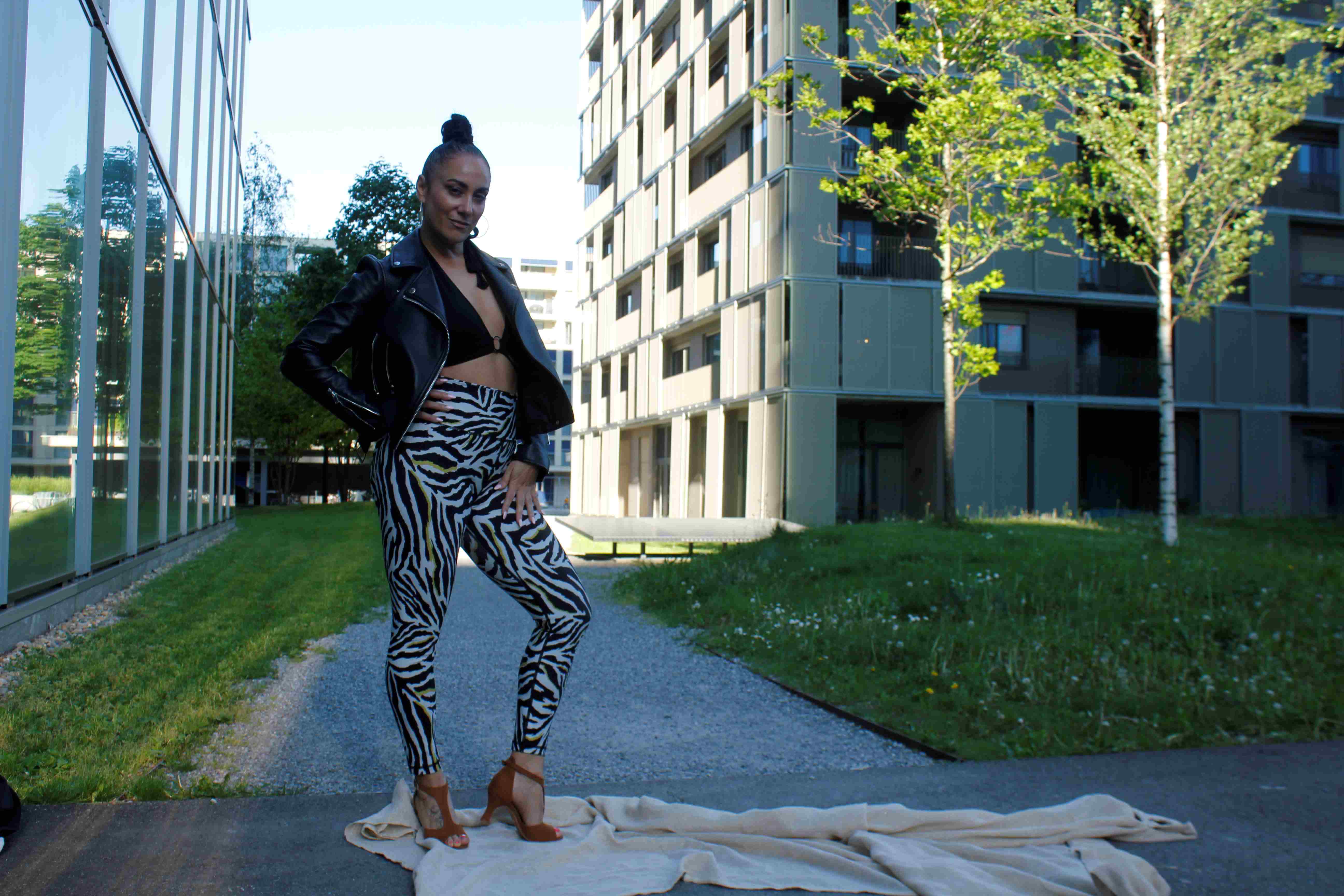 Frau in Hose mit Zebramuster und schwarzer Jacke posiert selbstbewusst auf einem Teppich vor modernen Gebäuden.