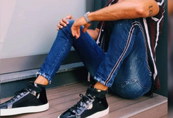 Person in Jeans und Turnschuhen sitzt mit gekreuzten Beinen auf einem Holzboden und trägt ein gestreiftes Hemd.
