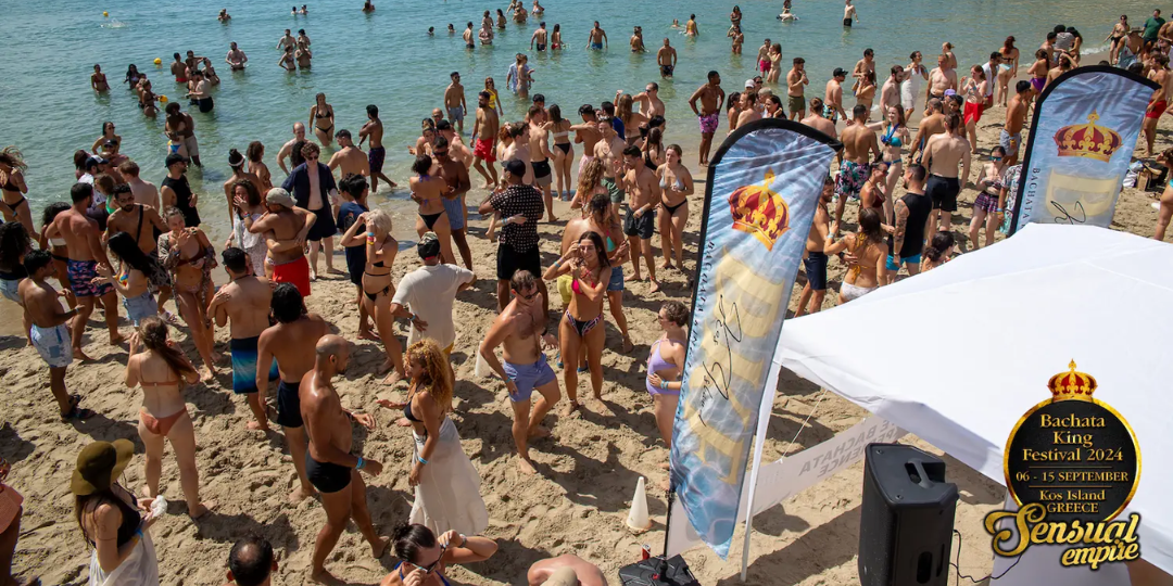 Eine tanzende und mischende Menschenmenge an einem sonnigen Strand während des Bachata King Festivals, mit Bannern und Menschen in Badekleidung.