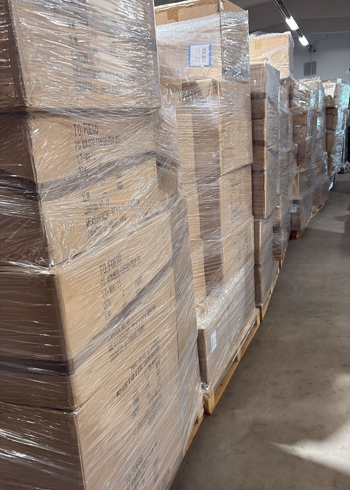 In einem Lagergang stehen reihenweise große, in Plastik verpackte Kartons auf Paletten.