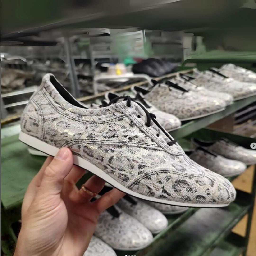 Eine Hand hält einen gemusterten Schuh, im Hintergrund stehen weitere ähnliche Schuhe auf einem Ständer.