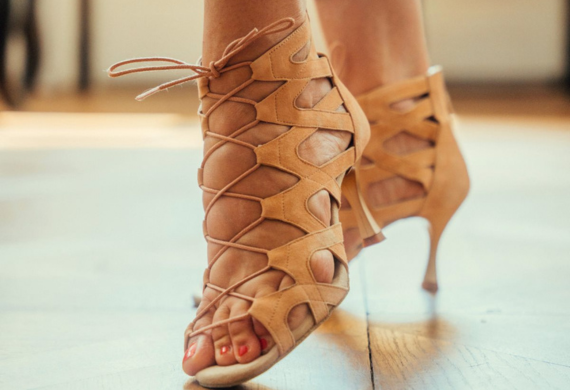 Nahaufnahme von Füßen in hellbraunen, hochhackigen Schnürsandalen auf einem Holzboden, mit lackierten Zehennägeln.