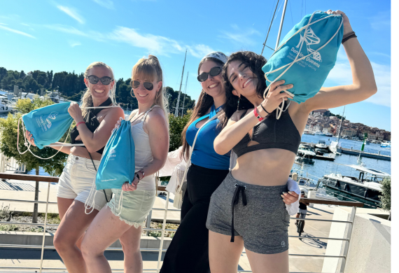 Vier lächelnde Frauen, die an einem sonnigen Tag mit blauen Kordeltaschen in der Nähe eines Jachthafens posieren.