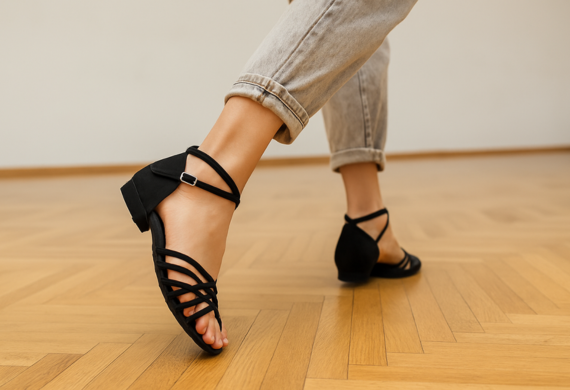 Person in schwarzen Riemchensandalen und hellen Jeans, die auf einem Holzboden geht.