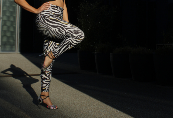 Person in Leggings und High Heels mit Zebramuster steht auf einem Bein in einem schattigen Bereich.
