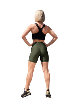 Eine Person in einem schwarzen Sport-BH und Lisminas 7009 Midi Short in Khaki steht mit den Händen in den Hüften und blickt abgewandt.