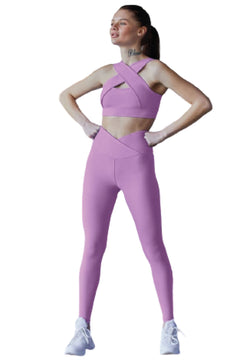 Eine Frau in einem rosa Outfit mit gekreuzten Trägern und weißen Schuhen steht selbstbewusst in Lisminas lila Leggings mit extra hoher Taille.