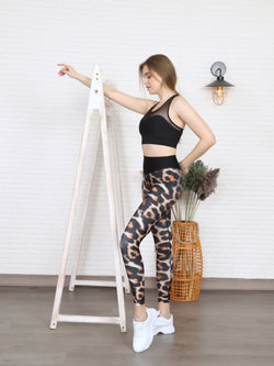 Eine Frau posiert neben einer Holzleiter und trägt Lisminas 1080 Extra High Waist Leopard Leggings und einen schwarzen Sport-BH.