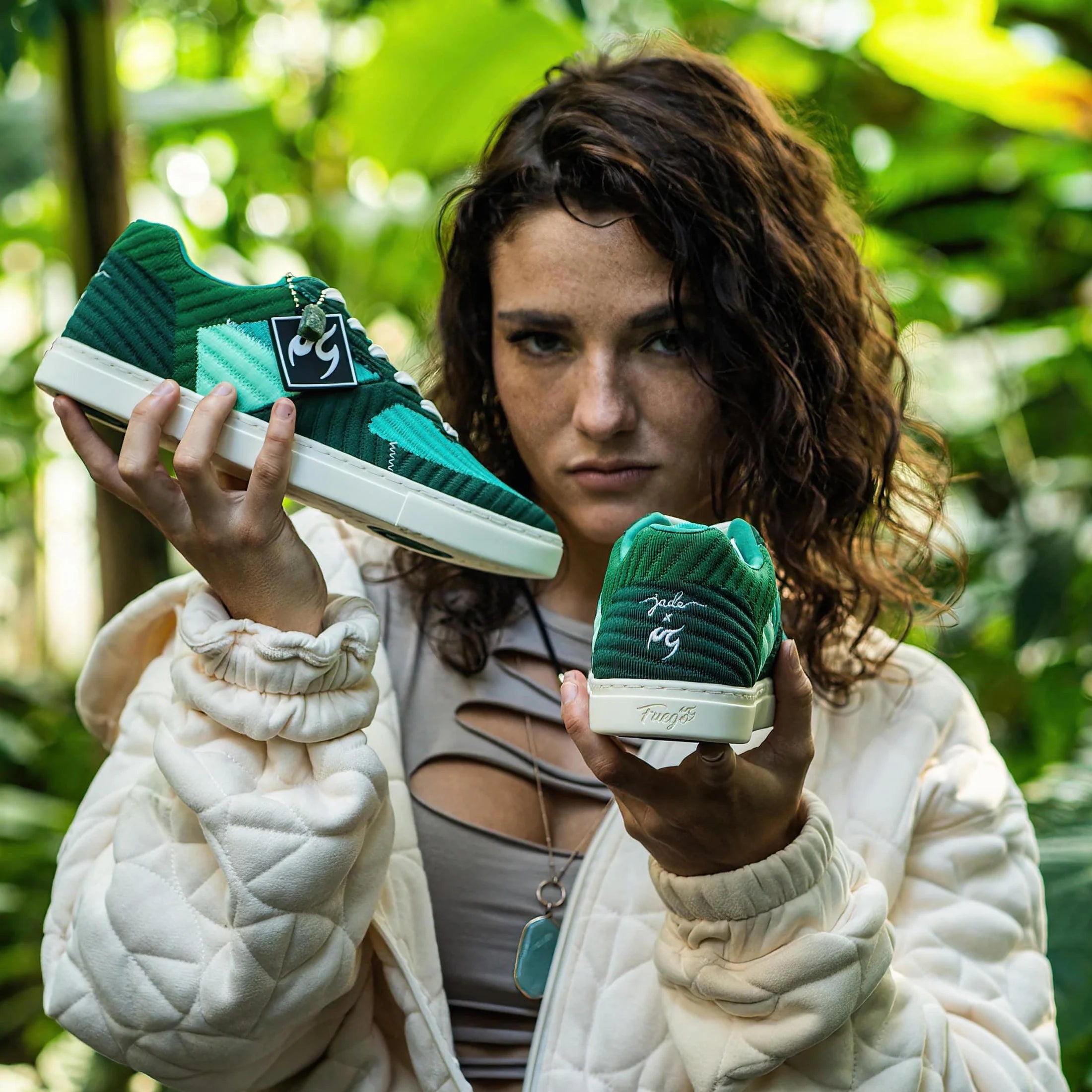 Jade Chynoweth hält in einer Steppjacke Fuego Jade Dance Sneakers von Fuego Dance Shoes in einer üppigen Dschungelkulisse.